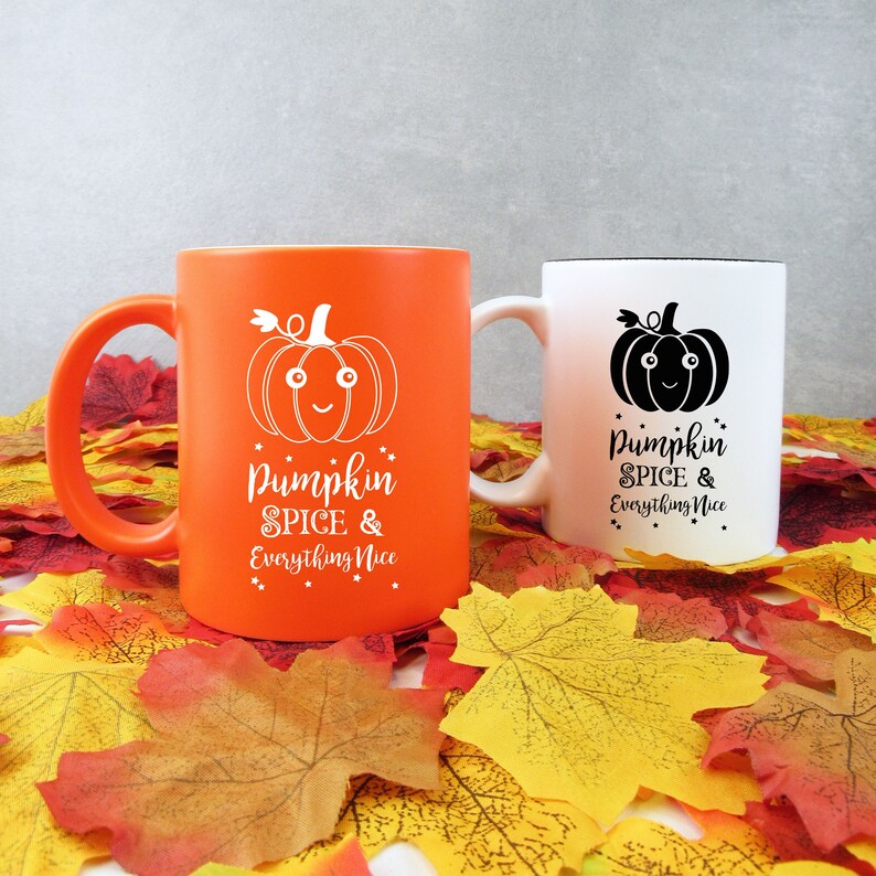 May include: Two ceramic mugs with fall-themed designs. One is orange with a white pumpkin graphic and text, the other is white with a black pumpkin graphic and text. The text on both mugs reads "Pumpkin Spice & Everything Nice."