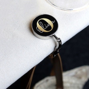 May include: A black and gold personalized bag hook with the name "Olivia" written in gold script on a black circle.