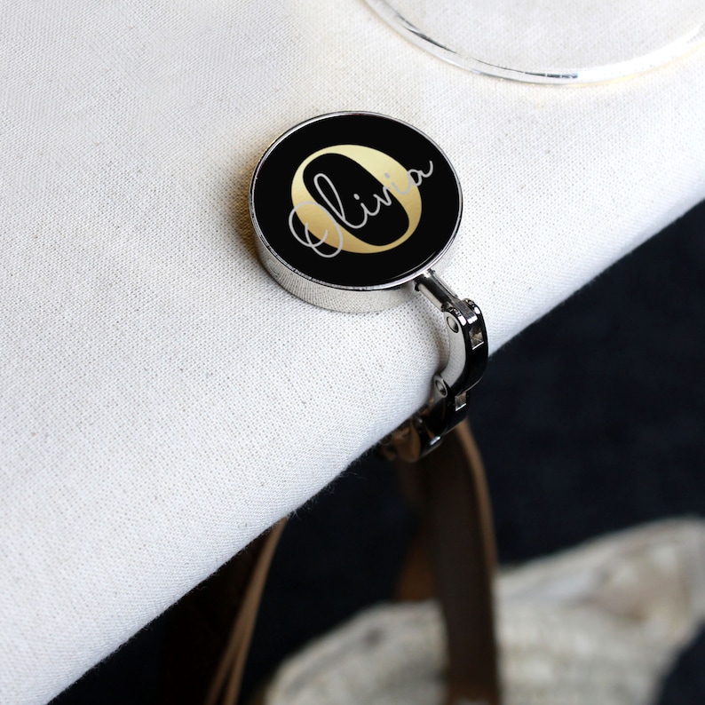 May include: A black and gold personalized bag hook with the name "Olivia" written in gold script on a black circle.