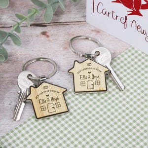 May include: Two wooden house-shaped keyrings with silver key rings and keys. The keyrings are engraved with "2025", "Ein Cartref Cyntaf", a heart, "Ellis & Lloyd", and a small house.