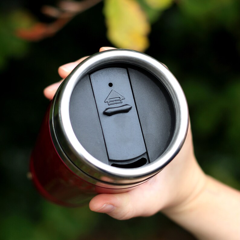 May include: A red travel mug with a stainless steel lid. The lid has a black push button with a symbol that looks like a house.