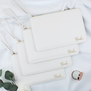 May include: Four white zippered pouches with gold handles and the word "Bride" written in gold script on each pouch.