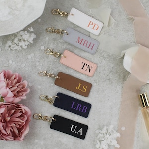 Op de afbeelding: Een verzameling gepersonaliseerde sleutelhangers in diverse kleuren: wit, grijs, roze, bruin, marineblauw en zwart. Elke sleutelhanger heeft initialen in een contrasterende kleur en een goudkleurige sluiting. De afbeelding bevat ook roze bloemen en een parfumflesje.