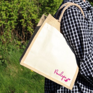 May include: A natural jute tote bag with a white canvas insert. The bag has a pink butterfly design and the name "Penelope" printed on the front.