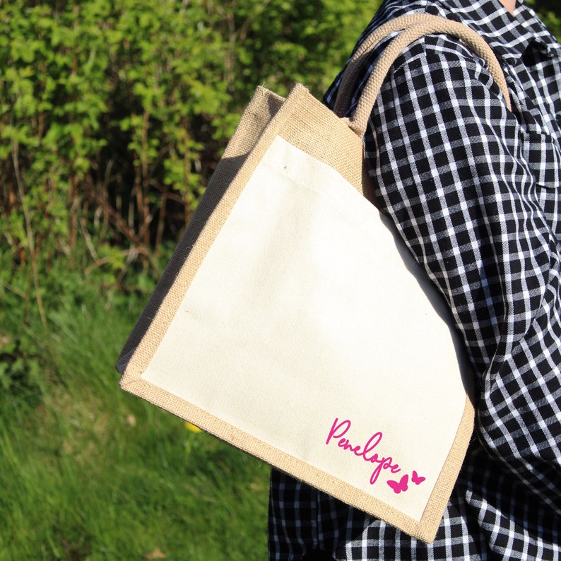 May include: A natural jute tote bag with a white canvas insert. The bag has a pink butterfly design and the name "Penelope" printed on the front.