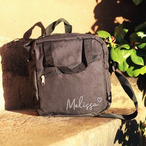 K&ouml;nnte beinhalten: Schwarze Canvas-Schultertasche mit Rei&szlig;verschlussfach und einem wei&szlig;en gestickten Namen "Melissa" mit einem Herz.