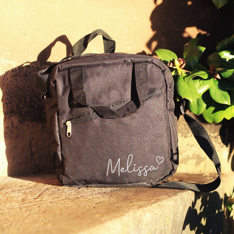 K&ouml;nnte beinhalten: Schwarze Canvas-Schultertasche mit Rei&szlig;verschlussfach und einem wei&szlig;en gestickten Namen "Melissa" mit einem Herz.