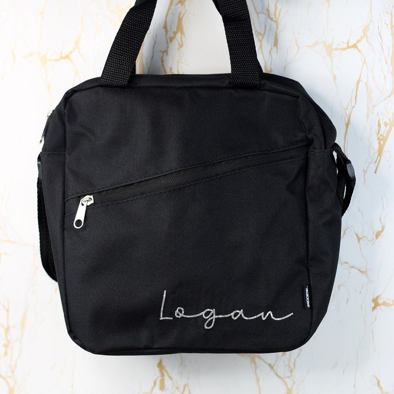 Pode incluir: Bolsa de almo&ccedil;o de lona preta com um bolso com z&iacute;per e um puxador de z&iacute;per prateado. A bolsa tem o nome "Logan" bordado em branco na frente.
