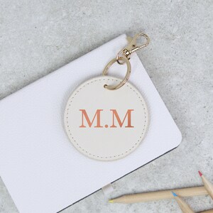May include: A white leather keychain with a gold key ring and a round cream-colored tag with the initials "M.M" in rose gold.