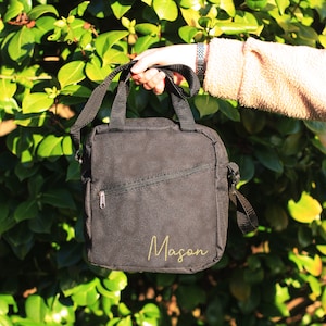 K&ouml;nnte beinhalten: Schwarze Canvas-Schultertasche mit Rei&szlig;verschlussfach und dem Namen "Mason" in goldenen Buchstaben.