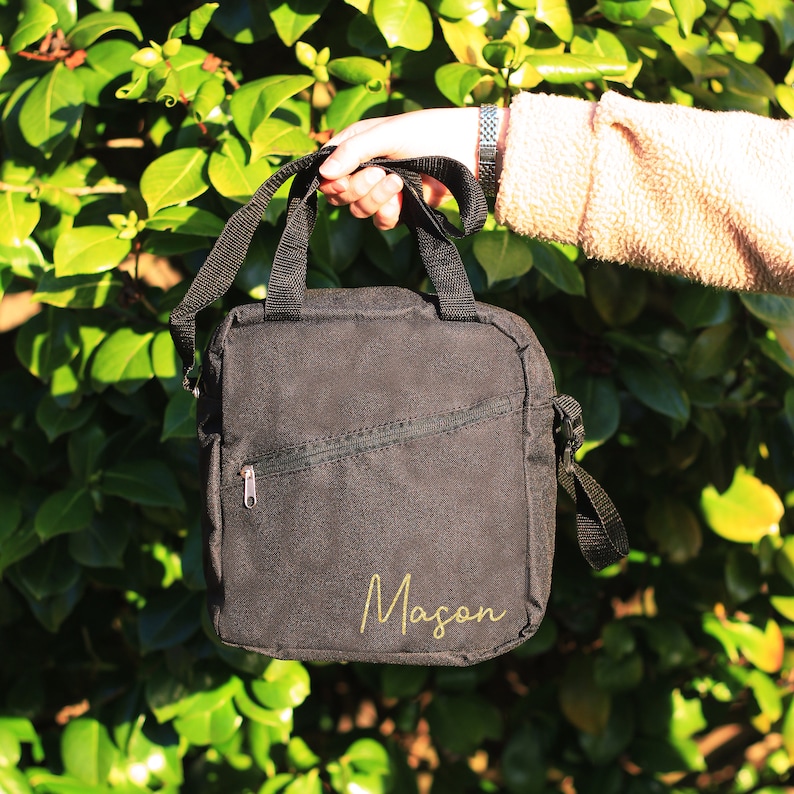 K&ouml;nnte beinhalten: Schwarze Canvas-Schultertasche mit Rei&szlig;verschlussfach und dem Namen "Mason" in goldenen Buchstaben.