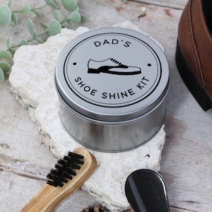 Könnte beinhalten: Ein silbernes Schuhputzset für Papa, mit dem Text "DAD'S SHOE SHINE KIT" um eine Schuhgrafik. Das Set enthält eine Bürste und einen Schuhanzieher. Ein brauner Lederschuh ist teilweise im Hintergrund zu sehen.