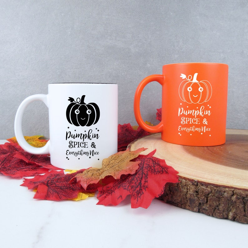 May include: Two ceramic mugs with fall-themed designs. One mug is white with a black pumpkin graphic and the text "Pumpkin Spice & Everything Nice." The other mug is orange with a white pumpkin graphic and the same text. The mugs are on a wooden surface with fall leaves.