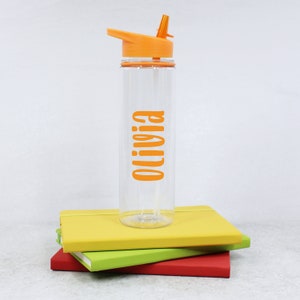 May include: Orange and clear plastic water bottle with a flip top lid and straw. The bottle has the name "Olivia" printed on it in orange.