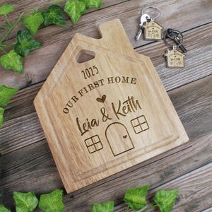 Op de afbeelding: Een houten snijplank in de vorm van een huis met de tekst "2025 Our First Home Leia & Keith" erop gegraveerd. De plank heeft een klein hartvormig uitsparing en een klein hart dat op de voorkant is gegraveerd.