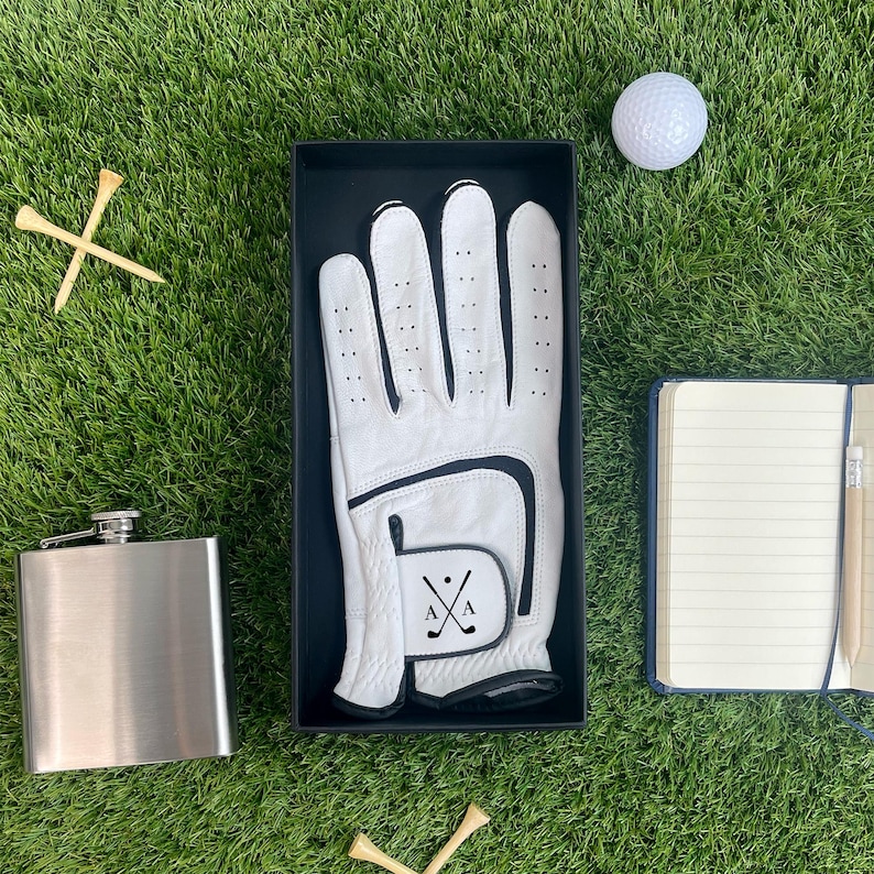 K&ouml;nnte beinhalten: Ein Golf-Geschenkset auf gr&uuml;nem Rasen. Das Set enth&auml;lt einen wei&szlig;en Golfhandschuh in einer schwarzen Box, einen Golfball, eine silberne Flasche, einen Notizblock mit Bleistift und h&ouml;lzerne Golf-Tees. Der Handschuh hat einen schwarzen Akzent.