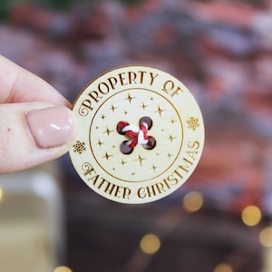 以下が含まれることがあります： 「PROPERTY OF FATHER CHRISTMAS」という文字が書かれた木製のボタン。ボタンには穴があり、赤と白の紐が通されています。ボタンには小さな星が飾られています。