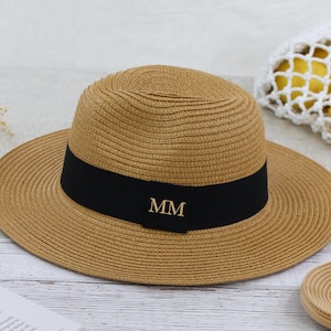 Può includere: Un cappello di paglia con una fascia nera e le iniziali dorate "MM" sul davanti. Il cappello è marrone e ha un'ampia tesa.