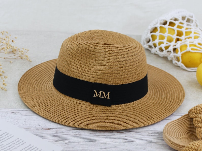 Peut inclure: Un chapeau de paille avec un bandeau noir et les initiales dor&eacute;es "MM" sur le devant. Le chapeau est brun et a une large bordure.