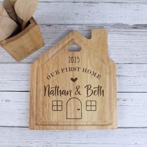 Op de afbeelding: Een houten snijplank in de vorm van een huis met de tekst "2025 Our First Home Nathan & Beth" erop gegraveerd.