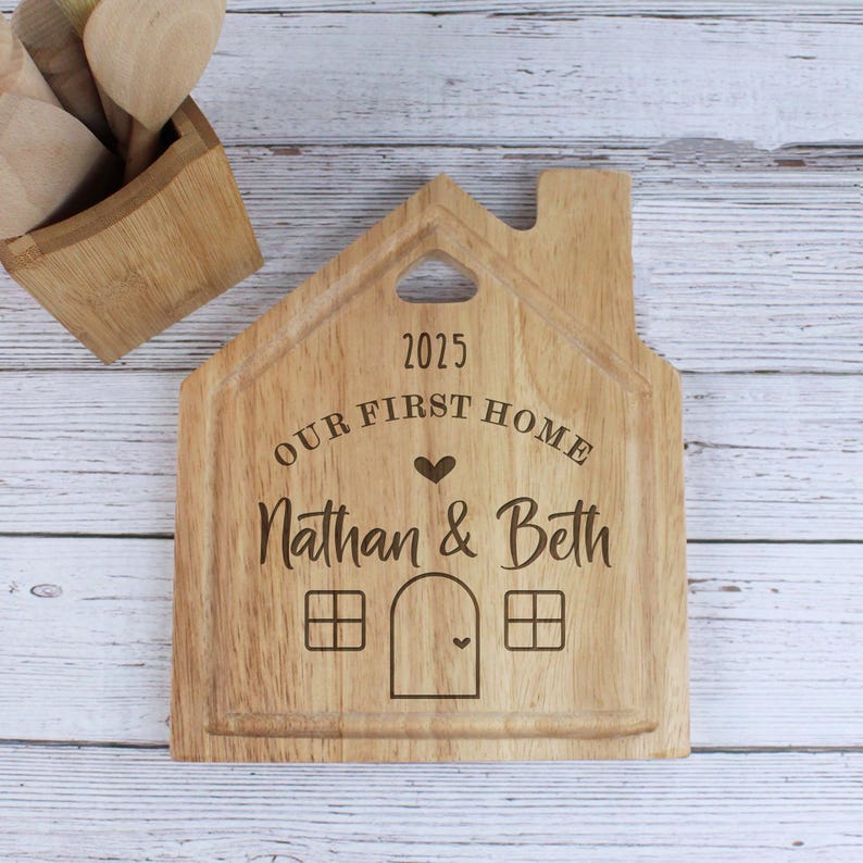 Op de afbeelding: Een houten snijplank in de vorm van een huis met de tekst "2025 Our First Home Nathan & Beth" erop gegraveerd.