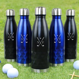 Puede incluir: Cinco botellas de agua aisladas en negro y azul con tapas plateadas. Cada botella tiene un diseño temático de golf con iniciales. Pelotas y tees de golf sobre hierba verde.
