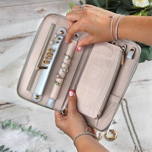 May include: A light grey jewellery organiser with a zip closure, holding various rings, earrings, and bracelets. The interior features a light pink lining and multiple compartments for storage. Gold-toned hardware accents the design.