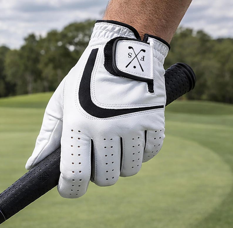 K&ouml;nnte beinhalten: Wei&szlig;er Golfhandschuh mit schwarzen Akzenten, gehalten an einem Golfschl&auml;ger. Der Handschuh hat ein schwarzes geschwungenes Design und einen kleinen Aufn&auml;her mit den Buchstaben "S" und "F", die mit einem Golfschl&auml;ger gekreuzt sind. Der Handschuh hat kleine Bel&uuml;ftungsl&ouml;cher.