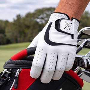 K&ouml;nnte beinhalten: Wei&szlig;er Golfhandschuh mit schwarzen Akzenten und einem kleinen Emblem mit gekreuzten Golfschl&auml;gern und den Buchstaben "D" und "E". Der Handschuh hat kleine Perforationen und liegt auf einer rot-schwarzen Golftasche.