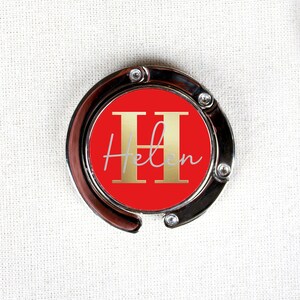May include: A silver-toned bag clip with a red circle featuring a gold letter "H" and the name "Helen" in white script.