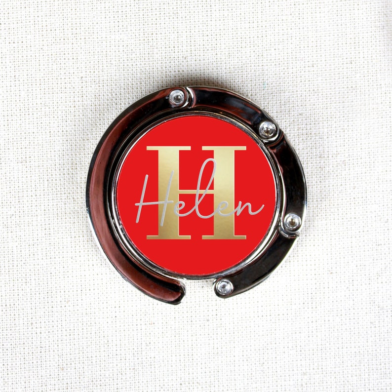 May include: A silver-toned bag clip with a red circle featuring a gold letter "H" and the name "Helen" in white script.