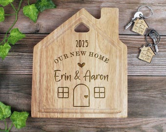 Könnte beinhalten: Ein hölzernes, hausförmiges Schneidebrett mit dem eingravierten Text "2025 Our New Home Erin & Aaron". Das Brett hat einen kleinen Herz-Ausschnitt und zwei Fenster.