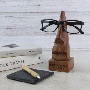 May include: Wooden stand shaped like a nose with a pair of black eyeglasses resting on it. The stand has a base with the text "Mr. Chadwick's Glasses" engraved on it.