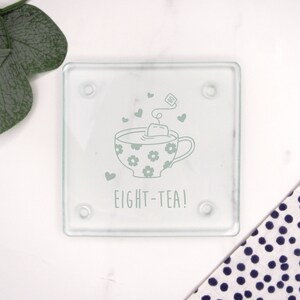 May include: A glass coaster with a white design of a teacup with a teabag and the text "EIGHT-TEA!"