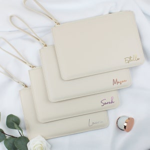 May include: Four beige zippered pouches with gold handles. Each pouch has a different name written on it in a different colour: Estelle, Mason, Sarah, and Laura.
