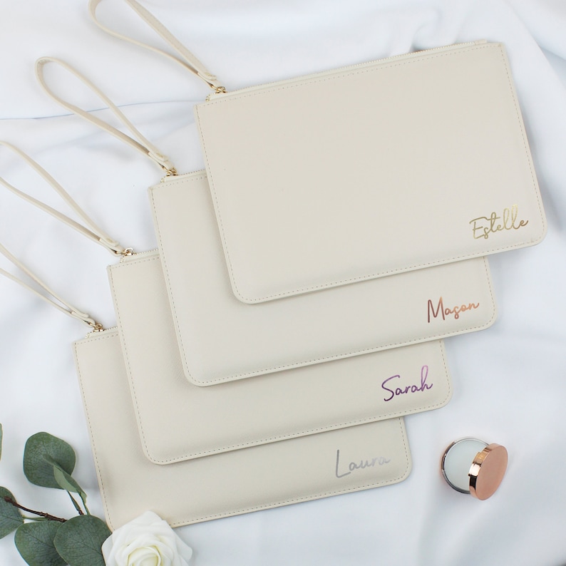 May include: Four beige zippered pouches with gold handles. Each pouch has a different name written on it in a different colour: Estelle, Mason, Sarah, and Laura.