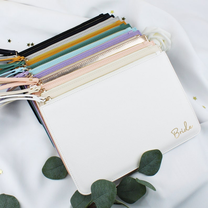 May include: A stack of zippered pouches in various colours, including black, gold, green, blue, and pink. The top pouch is white with the word "Bride" written in gold lettering.