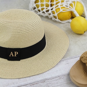 Peut inclure: Un chapeau de paille beige avec un ruban noir et les initiales dor&eacute;es "AP" sur le devant. Le chapeau est pos&eacute; sur une surface en bois blanc.