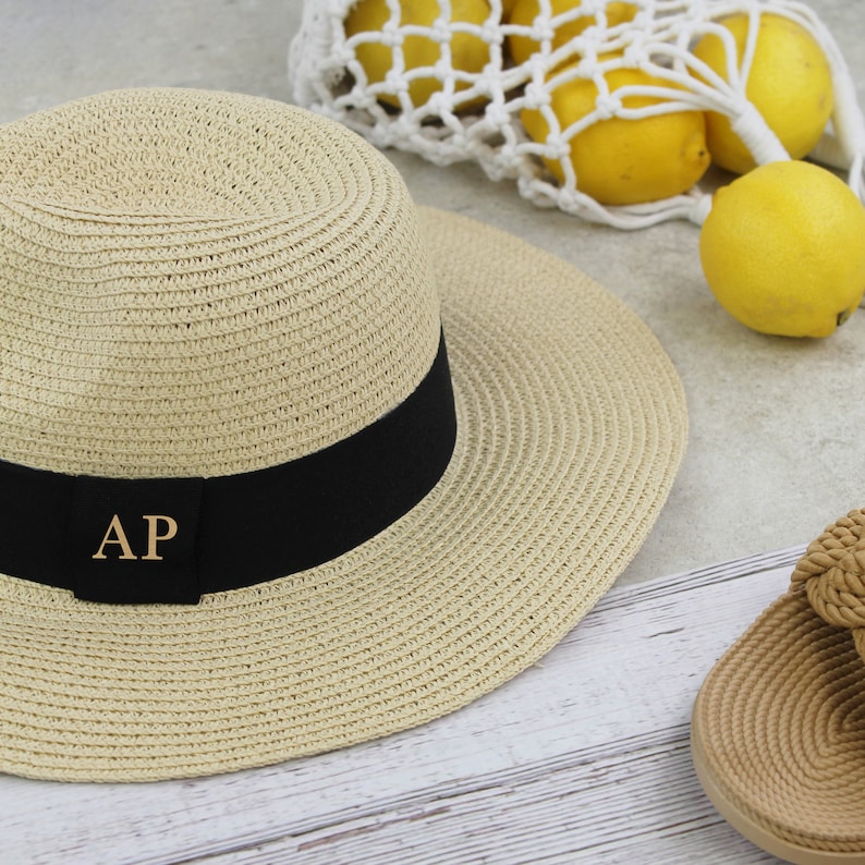Peut inclure: Un chapeau de paille beige avec un ruban noir et les initiales dor&eacute;es "AP" sur le devant. Le chapeau est pos&eacute; sur une surface en bois blanc.
