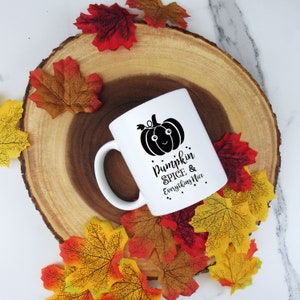 May include: White ceramic mug with a black pumpkin graphic and the text "Pumpkin Spice & Everything Nice." The mug is surrounded by colorful fall leaves and rests on a wooden slice.