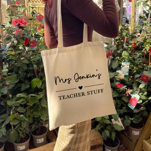 Op de afbeelding: Een crèmekleurige draagtas met de tekst "Mrs Jenkins's TEACHER STUFF" in zwart schrift. De tas wordt vastgehouden tegen een achtergrond van groene planten en rode bloemen.