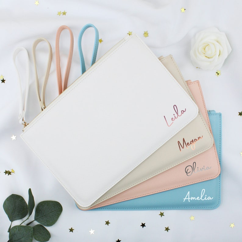May include: Four personalised zippered pouches in different colours: white, beige, pink, and blue. Each pouch has a different name written in gold lettering: Leila, Megan, Olivia, and Amelia.