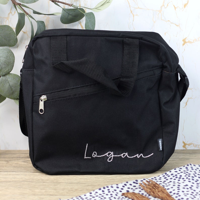Pode incluir: Bolsa de ombro de lona preta com um bolso com z&iacute;per e um nome bordado em rosa "Logan".