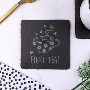 May include: Black slate coaster with a white outline of a teacup with a teabag and the text "EIGHT-TEA!"