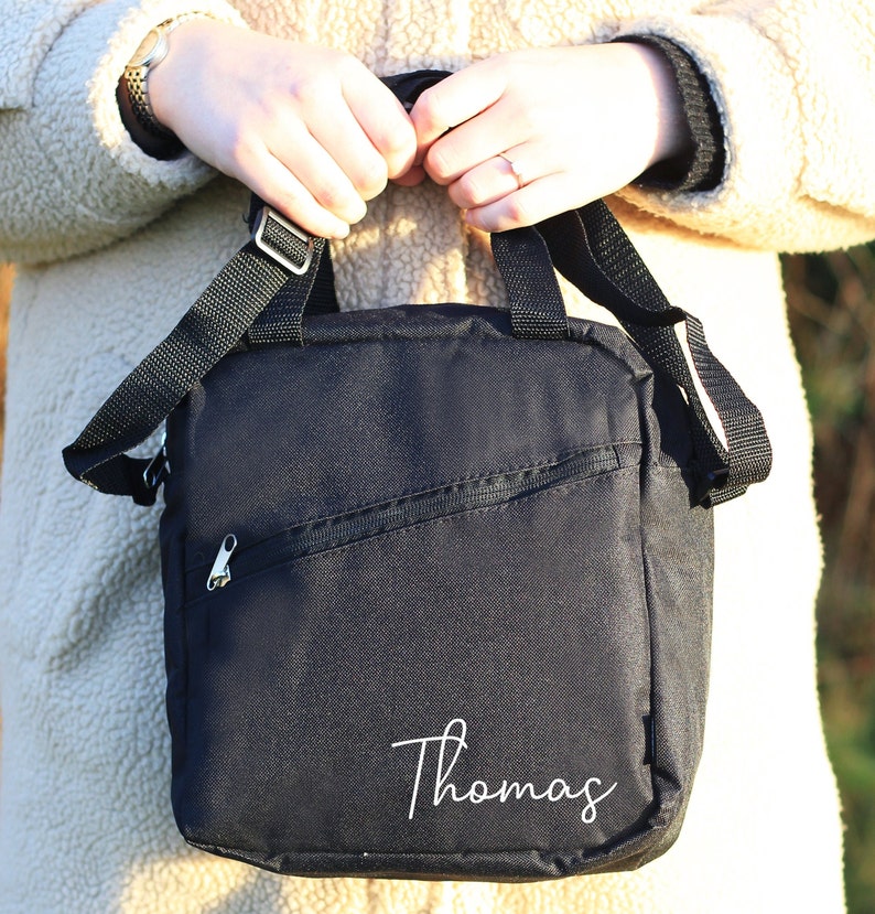 K&ouml;nnte beinhalten: Schwarze Umh&auml;ngetasche mit Rei&szlig;verschlussfach und einem wei&szlig;en gestickten Namen "Thomas".
