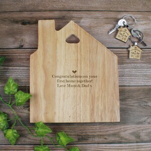 Op de afbeelding: Houten snijplank in de vorm van een huis met een hartvormige uitsparing en de gegraveerde boodschap "Gefeliciteerd met jullie eerste huis samen! Liefs, Mama & Papa x".