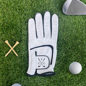 K&ouml;nnte beinhalten: Wei&szlig;er Golfhandschuh mit schwarzem Besatz und den Initialen "CG" mit gekreuzten Golfschl&auml;gern. Zwei Golf-Tees aus Holz und ein Golfball sind ebenfalls auf dem gr&uuml;nen Gras zu sehen, zusammen mit einem Golfschl&auml;ger.
