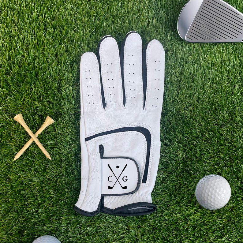 K&ouml;nnte beinhalten: Wei&szlig;er Golfhandschuh mit schwarzem Besatz und den Initialen "CG" mit gekreuzten Golfschl&auml;gern. Zwei Golf-Tees aus Holz und ein Golfball sind ebenfalls auf dem gr&uuml;nen Gras zu sehen, zusammen mit einem Golfschl&auml;ger.