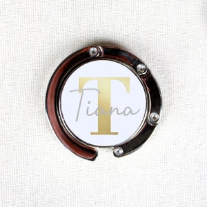 May include: A silver-toned bag hook with a white circle featuring a gold letter "T" and the name "Tiana" in a cursive font.