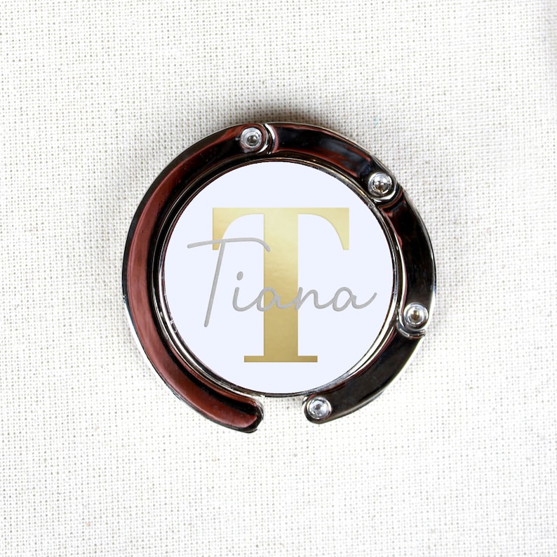 May include: A silver-toned bag hook with a white circle featuring a gold letter "T" and the name "Tiana" in a cursive font.